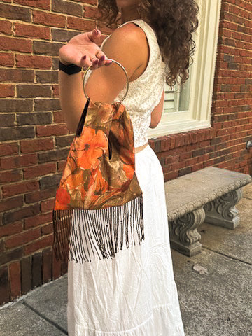 A woman in a white lace dress holds up a fringe bag with a rust-orange floral print and black fringe trim, standing outdoors against a red brick wall.