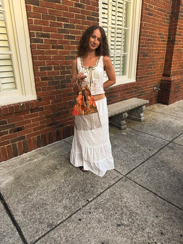 A woman stands on a stone sidewalk in front of a red brick building, wearing a white sleeveless bohemian-style maxi dress with a tie neckline and fringe detail. She is holding a patterned orange and brown handbag.