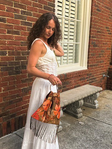A woman in a white lace top and white skirt holds a brown and orange patterned fringed handbag while standing outdoors against a red brick wall next to a stone bench.
