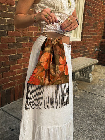 A person outdoors wearing a white skirt and holding a small top-handle bag with a brown and orange leaf print and long black fringe.