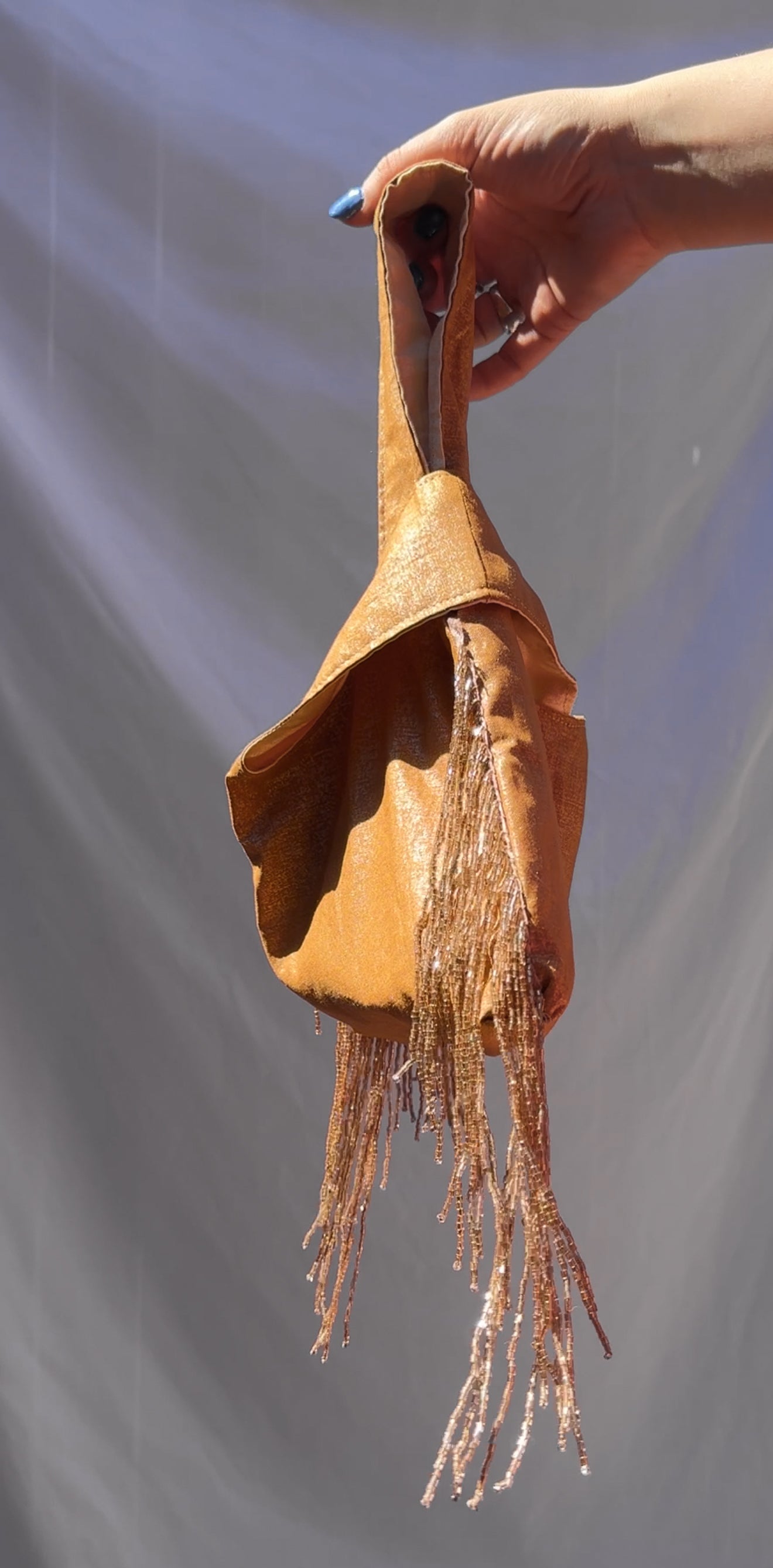 gold fabric bag with beaded fringes held by a hand against a gray background
