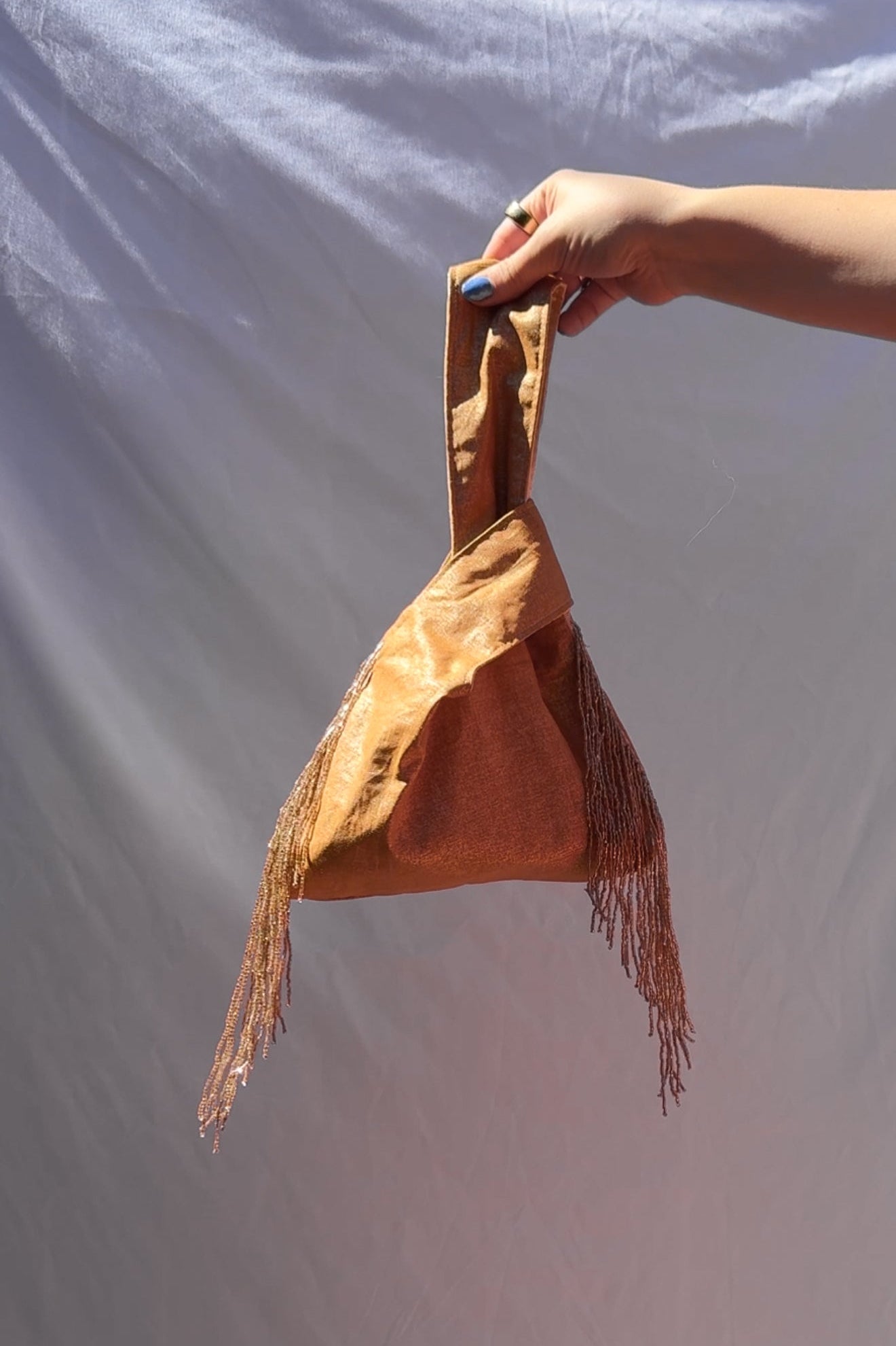 Hand holding a gold handbag with fringes against a plain background