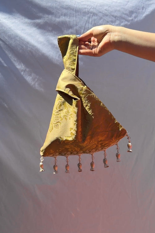 Hand holding a gold fabric item with tassels against a light blue background