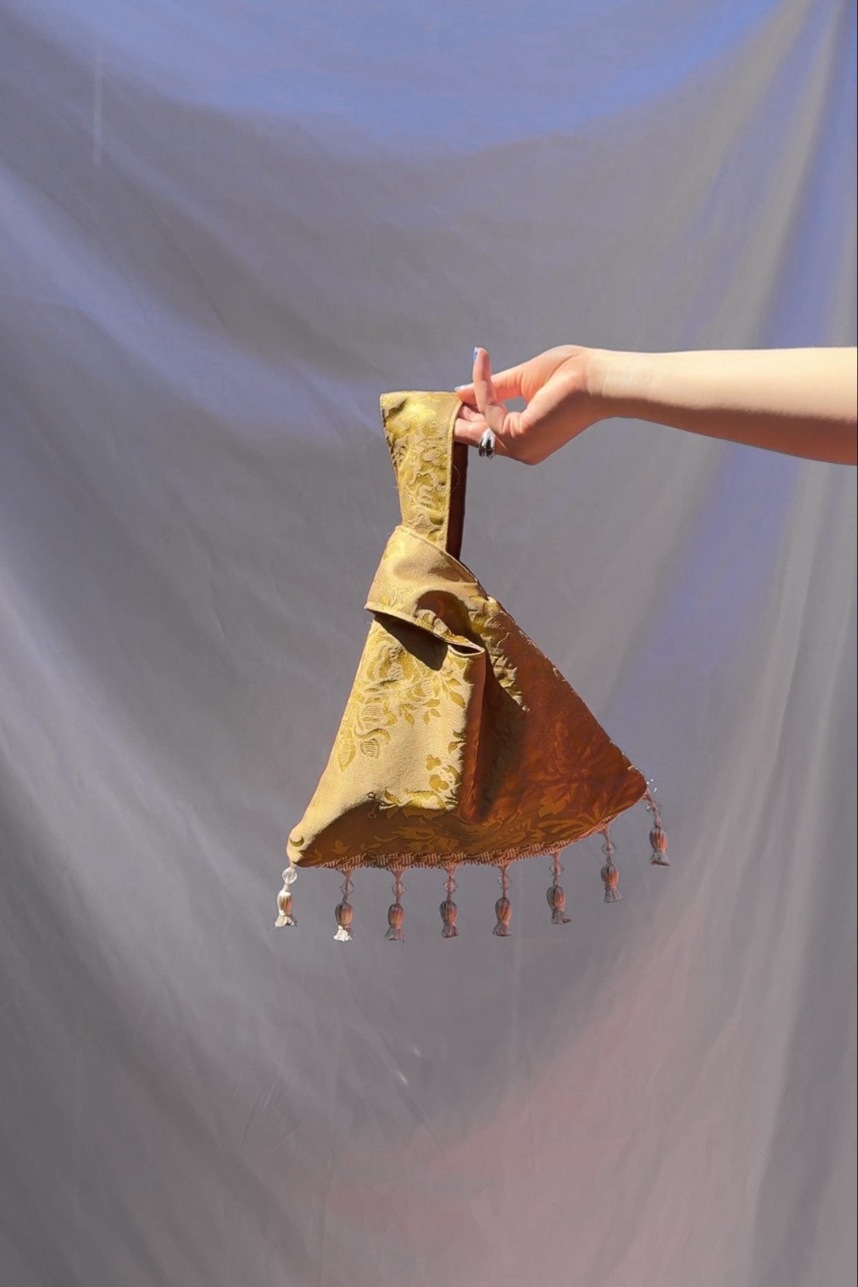 Gold handbag with tassels held by a hand against a plain background