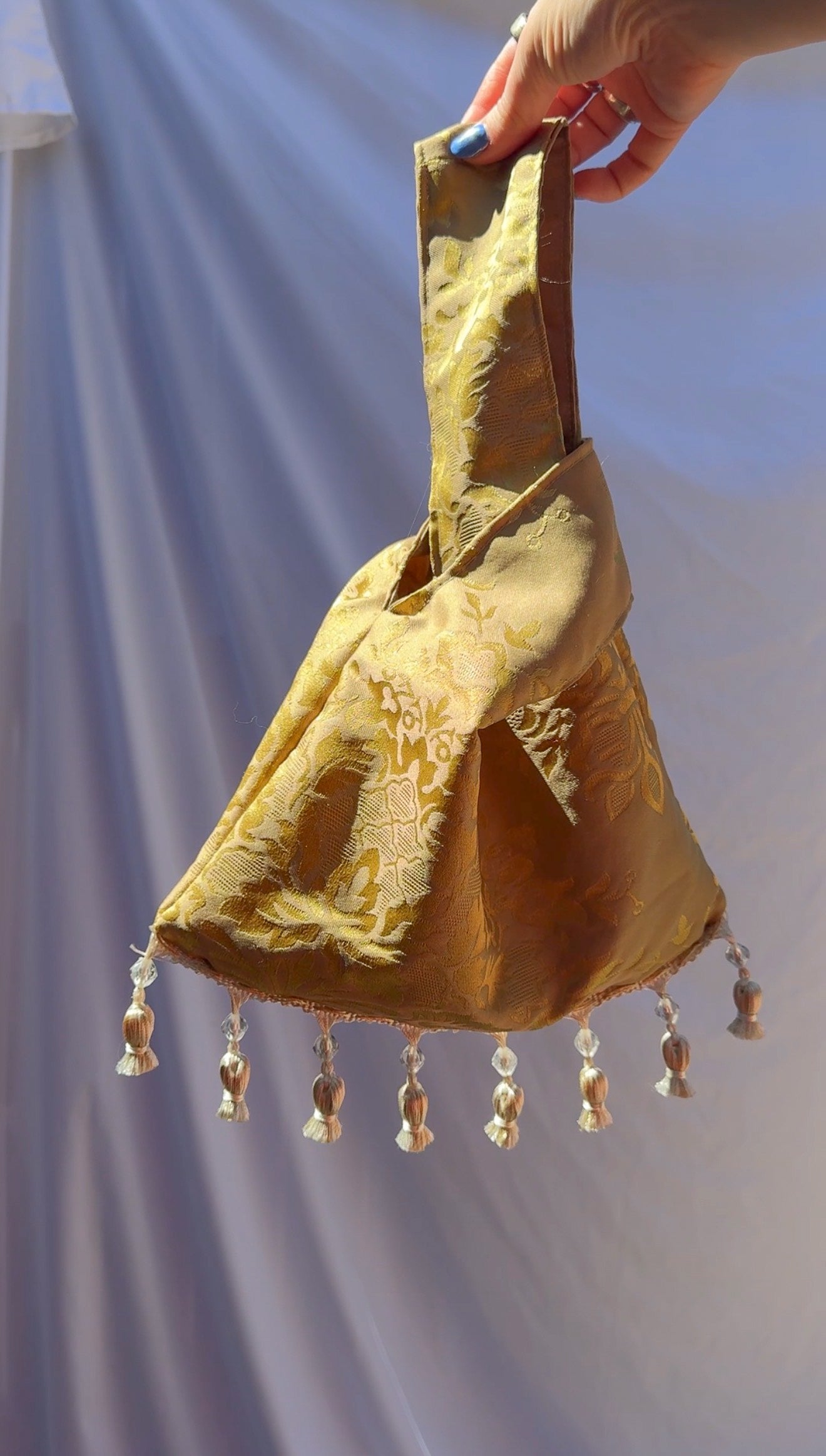 Hand holding a yellow fabric bag with tassels against a light blue curtain background