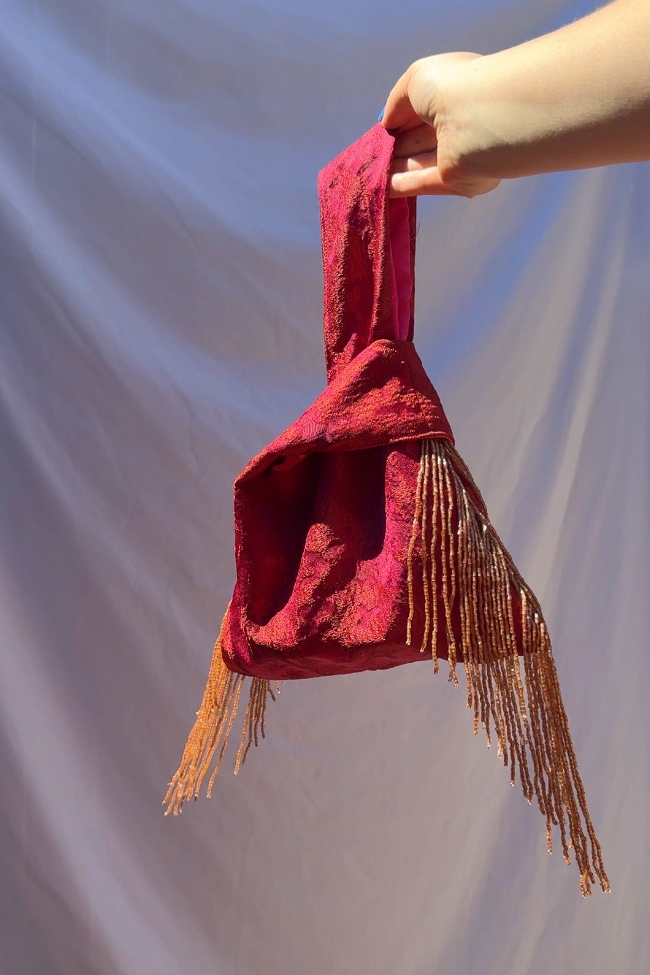 Fuchsia fabric bag with fringes held by a hand against a plain background