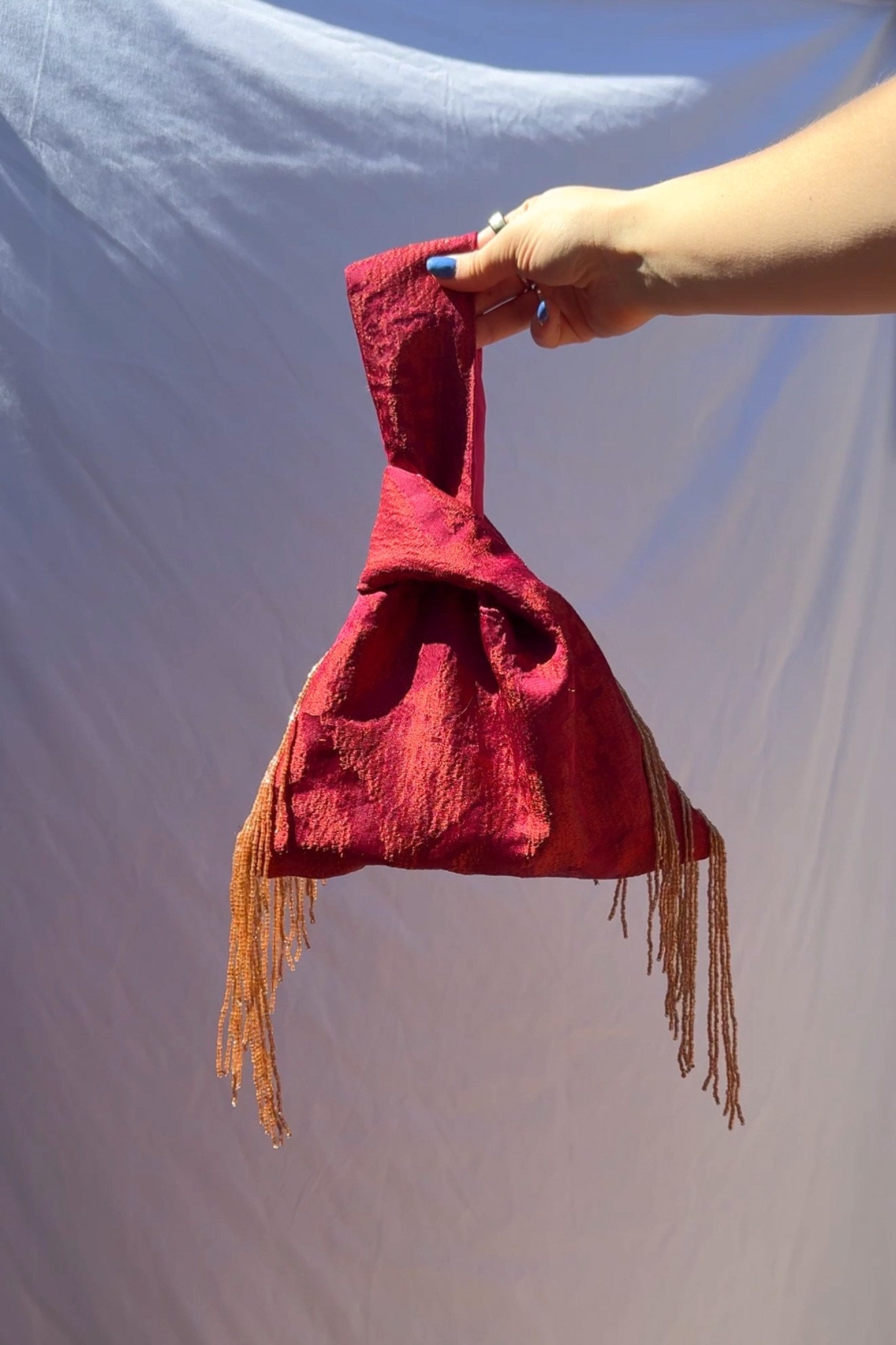 Fushcia fabric bag with fringes held by a hand against a plain background