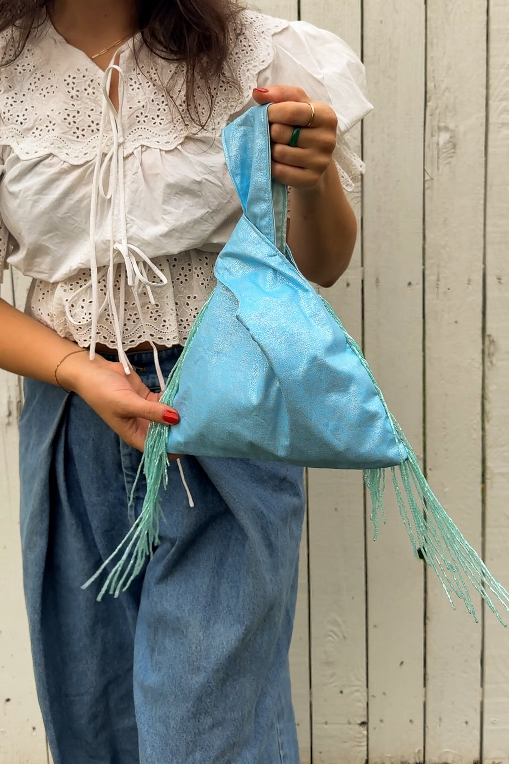 A person wearing a white lace-trim blouse and blue jeans holds a light blue fringed shoulder bag against a white wooden plank wall.