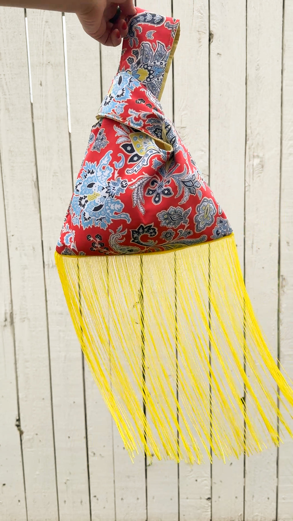 A hand holds a red patterned fabric shoulder bag with a bright yellow fringe bottom, set against a white wooden plank background.